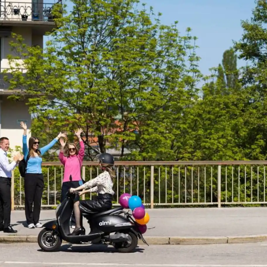 kvinna med hjälm kör moped och 3 personer står och vinkar när hon åker förbi.